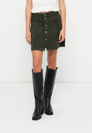 Femme portant une jupe en velours côtelé vert foncé avec une patte de boutonnage devant et de grandes poches, un top court blanc et des bottes noires montantes à talons.