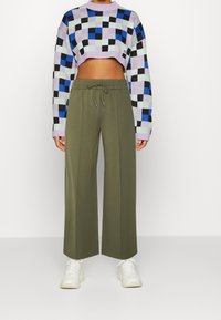 Persoon in een gekropte, meerkleurige geblokte trui, olijfgroene wijde broek met taillekoord en witte chunky sneakers.