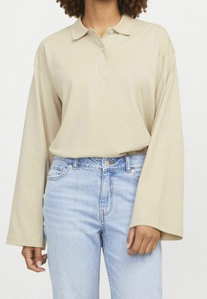 Persoon die een beige langarmig poloshirt draagt, ingestopt in lichtblauwe high-waisted jeans, staand tegen een effen achtergrond.