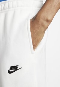 Vita Nike-sweatpants med elastisk midja, sidoficka och svart broderad logga på låret. Slät tygtextur.