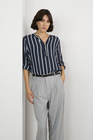 Blusa a righe navy con scollatura a V e dettagli con bottoni; pantaloni grigi a vita alta con pence; cintura nera in evidenza. Modella appoggiata a un muro.