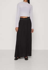 Lichtpaarse top met lange mouwen, geribbelde textuur en versierde randen, gecombineerd met een zwarte maxirok met elastische tailleband. Zwarte platte schoenen met studs.