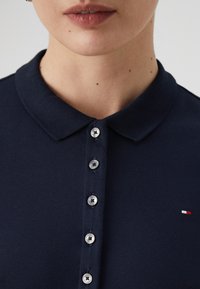 Granatowa koszulka polo z fakturowanej tkaniny, z płaskim kołnierzykiem, sześcioma srebrnymi guzikami i małym logo w kolorach czerwonym, białym i niebieskim.