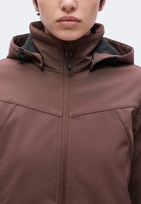 Personne portant une veste marron zippée avec un col montant et une capuche attachée sur un fond clair uni.