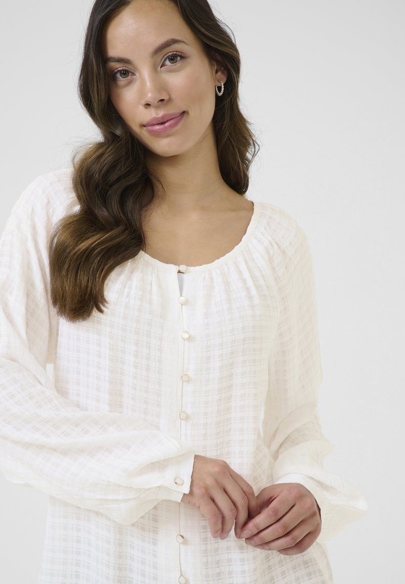 White, textured blouse with a round neckline and button closure. Features long sleeves and subtle checkered pattern.