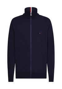 Sudadera de cremallera azul marino con cuello alto, puños y dobladillo acanalados. Presenta un pequeño logo bordado en el pecho y un acento a rayas en el cuello.