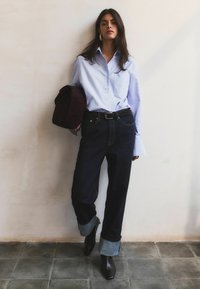Mujer con camisa azul claro abotonada, jeans oscuros con puños, botas negras, sosteniendo una prenda oscura doblada, de pie contra una pared pálida y suelo de baldosas.