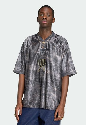 Joven con una camiseta deportiva holgada de color gris con patrón y el número "94" en dorado en el frente, combinada con pantalones azules y joyas doradas.