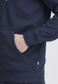 Marineblauwe sweatshirt met een voorzak, ribgebreide mouwen en een zijkraag. Materiaal lijkt glad met een zachte textuur.