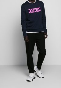 Person som har på sig en svart sweatshirt med rosa spegelvänd text "HUGO", svarta byxor, en vit T-shirt under, och vita sneakers mot en grå bakgrund.