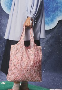 Asmuo laiko rožinės spalvos krepšį su gėlių raštu ir animacinių personažų piešiniais, dėvi šviesiai mėlyną marškinius ir tamsią sijoną, fone matomas žvaigždėtas dangus.