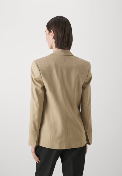 Beige maßgeschneiderte Blazer mit strukturiertem Design, scharfen Revers und figurbetonter Silhouette. Das Material wirkt glatt mit subtiler Textur; einzelner Schlitz hinten.