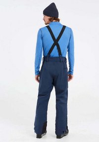 Jūriniai sniego kelnes su reguliuojamais juodais petnešomis, pagamintos iš patvarios medžiagos, su sustiprintais apatiniais kojų galiukais ir laisvu siluetu, dėvimos ant mėlynos viršutinės dalies.