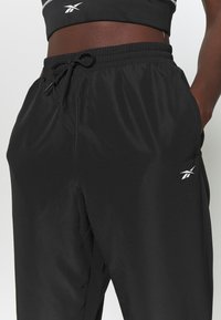 Černé sportovní kalhoty z lehkého materiálu, s elastickým pasem s tkaničkou a malým bílým logem na levé noze.