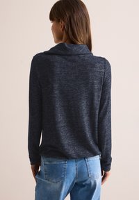 Maglione blu navy a maniche lunghe con collo a cowl, caratterizzato da un tessuto testurizzato e una vestibilità morbida, abbinato a jeans di denim azzurri.