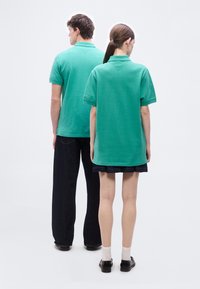 Δύο άτομα με την πλάτη γυρισμένη, φορούν ίδια πράσινα πόλο μπλουζάκια· το ένα φοράει μαύρο παντελόνι με φαρδιά πόδια, το άλλο μαύρη φούστα, άσπρες κάλτσες και μαύρα παπούτσια.