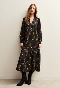 Vrouw die een lange zwarte jurk draagt met een gele bloemenprint en ruches, gecombineerd met zwarte laarzen met hak en gelaagde gouden kettingen.