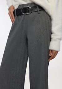 Zbliżenie na osobę ubraną w szare spodnie w jodełkę z czarnym paskiem i srebrną klamrą, w połączeniu z białym, puszystym swetrem z długim rękawem.