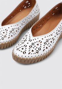 Zapatos blancos de cuero tipo slip-on con un patrón floral recortado, que presentan una suela de cuña de goma marrón y un acabado de costura.