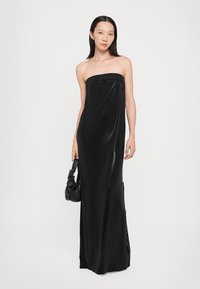 Black strapless evening gown in a slim fit, featuring a smooth, shiny texture and side slit. Model holding a textured black handbag.