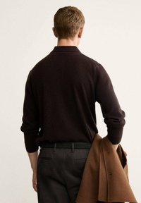 Maglione marrone scuro con colletto polo, maniche lunghe e un tessuto strutturato, abbinato a pantaloni scuri e una giacca marrone appoggiata sull'avambraccio.
