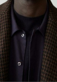 Hnědý houndstooth blazer se strukturovanými klopami přes tmavou košili s knoflíky. Obsahuje stříbrné knoflíky a měkký strukturovaný černý rolák.