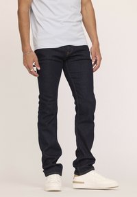 Jean en denim foncé avec une coupe slim, dotés d'une taille standard, de coutures discrètes et d'un design à jambe droite, associés à des baskets blanches.