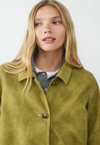 Oliwkowa skórzana kurtka z wysokim kołnierzem, zapięciem na jeden guzik i widocznymi detalami szycia, noszona na szarym podkoszulku.