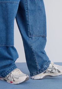 Denim bikses vidēji zilā krāsā, brīvs piegriezums ar taisnu kāju; kombinētas ar baltām sporta kedām ar pelēkiem akcentiem un teksturētām zolitēm.