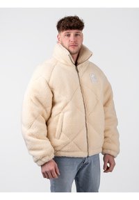 Chaqueta de forro polar acolchada en color crema, con un cuello alto, cierre de cremallera y dos bolsillos laterales. La tela tiene un acabado suave y texturizado.