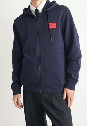 Uomo che indossa una felpa con cappuccio blu navy con patch rossa "HUGO", camicia bianca, cravatta nera e pantaloni neri, in piedi con le mani rilassate.