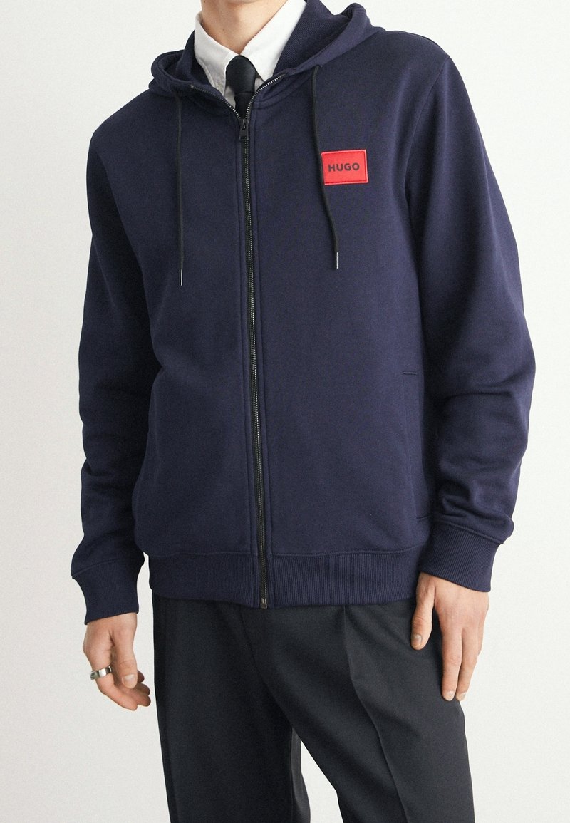 Hombre vestido con una sudadera con cremallera azul marino y parches de logo rojo "HUGO", camisa blanca, corbata negra y pantalones negros, de pie con las manos relajadas.