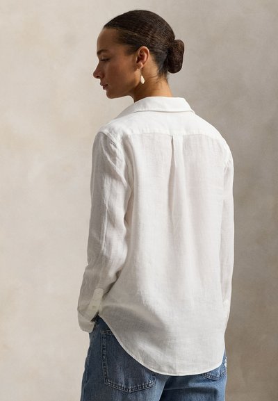 Λευκό πουκάμισο από λινό με άνετη εφαρμογή, γιακά, πτυχές στην πλάτη και μανσέτες με κουμπιά, σε συνδυασμό με μπλε τζιν παντελόνι. Απλός και υφασμάτινος σχεδιασμός.
