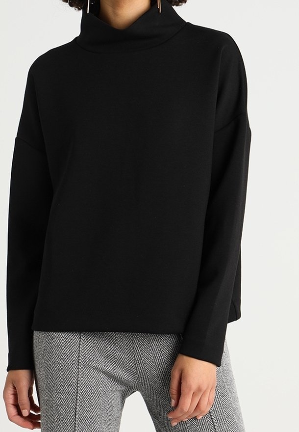 Personne portant un pull noir à col roulé manches longues et un pantalon gris à motif chevrons, debout devant un fond uni.