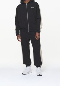 Hombre que lleva una sudadera con cremallera negra y pantalones deportivos negros a juego con rayas laterales beige y zapatillas blancas, de pie sobre un fondo blanco.