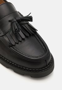 Must musta nahk loafer, millel on lame tald, dekoratiivsed franged ja tutid. Sile tekstuur koos kontrastsete õmblustega ääres.