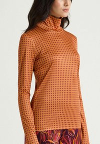 Col roulé à manches longues de couleur orange chaud avec des pois violets, texture lisse, coupe ajustée et col haut. Un imprimé floral apparaît en bas.