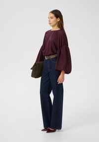 Dyp burgunder, flytende bluse med vide ermer, paret med høyt liv, mørkeblå denimjeans. Modellen holder en mørkegrønn veske.