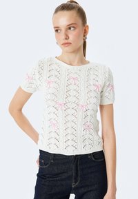 Hvid strikket kortærmet sweater med hulmønster, der har lyserøde sløjfedetaljer på ærmerne og en ribbet kant.