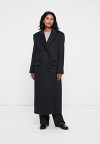 Cappotto lungo doppiopetto in lana grigia scura con rever strutturati, due tasche anteriori e silhouette dritta. Indossato sopra un top in maglia leggera.