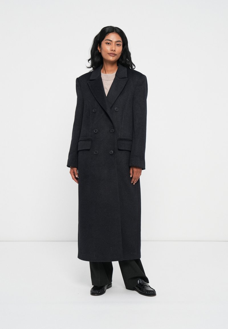 Cappotto lungo doppiopetto in lana grigia scura con rever strutturati, due tasche anteriori e silhouette dritta. Indossato sopra un top in maglia leggera.