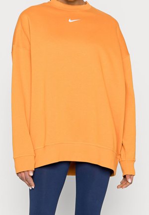 Personne portant un sweat-shirt Nike oversize orange vif à manches longues et des leggings bleu marine, debout devant un fond uni.