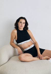 Crni sportski grudnjak s bijelim Calvin Klein logotipom na trakama, uparen s crnim kratkim hlačama, sjedi na teksturiranoj, svijetloj ležaljci.