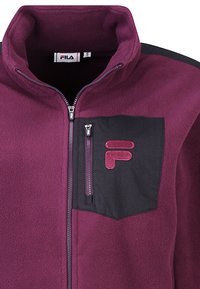 Chaqueta de forro polar en morado oscuro con cuello alto, cremallera completa y un bolsillo en el pecho negro que presenta un logotipo rojo. Textura suave y material duradero.