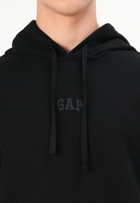 Black hoodie featuring a kangaroo pocket, adjustable drawstrings, and an embossed "GAP" logo on the chest in matching dark thread.