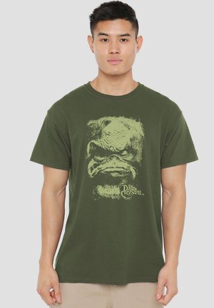 Young man wearing olive green t-shirt with a distressed graphic of a creature's face and "The Dark Crystal" text on the front.