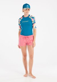 Teal rash guard s multicolor vzorovanými rukávmi a logom 'protest'; v kombinácii s ružovými plážovými šortkami. Model má na hlave modrú čiapku.
