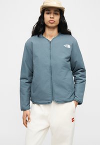 Νεαρή γυναίκα που φοράει μπεζ καπέλο, μπλε ζακέτα με φερμουάρ και λευκό παντελόνι με λο γότυπα The North Face στη ζακέτα και το παντελόνι.