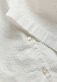 Λευκό ύφασμα με διακριτικό γεωμετρικό υφασμάτινο σχέδιο και δύο μικρά λευκά κουμπιά σε διπλωμένο τμήμα.