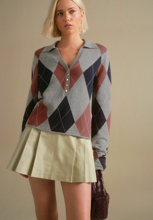 Grauer Argyle-Pullover mit roten und marineblauen Rauten, Knopfblende, kombiniert mit einem gefalteten hellgrünen Rock. Strukturiertes Gewebe, lässiges Design.